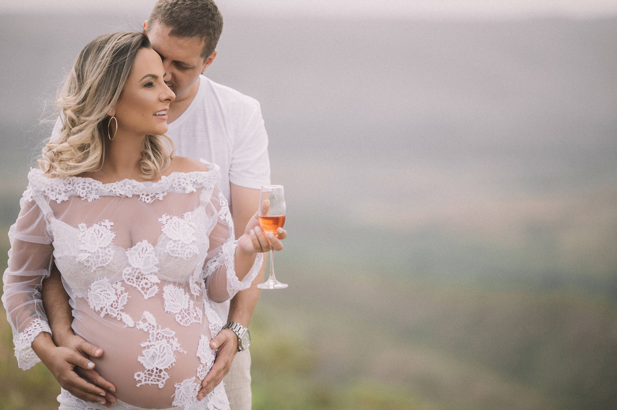 Gestante segurando uma taça de champagne abraçada com seu marido. Clicado pelo fotógrafo de gestantes Rafael Ohana em Brasília-DF