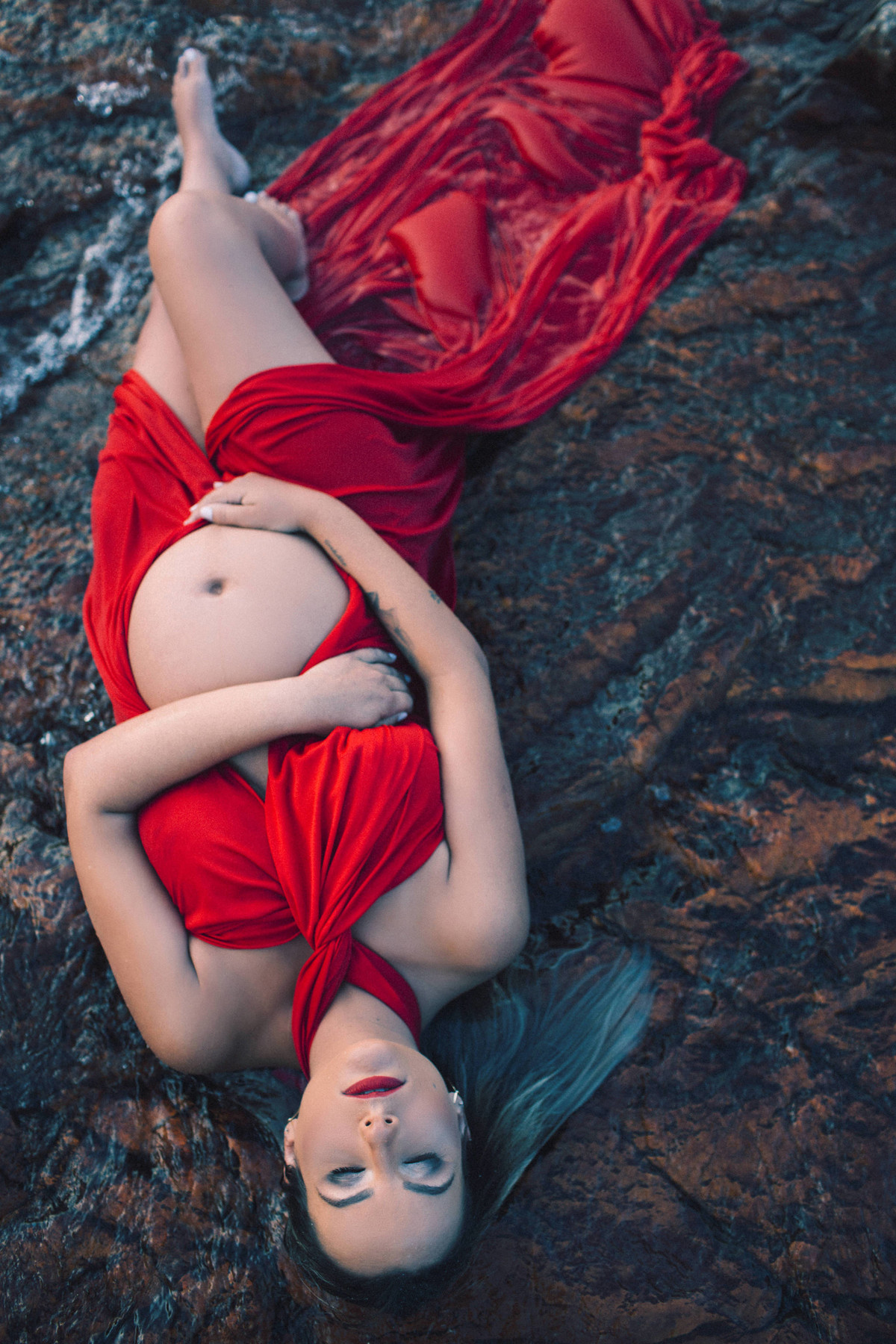 Grávida deitada nas pedras do rio usando um vestido vermelho. Clicado pelo fotógrafo de gestantes Rafael Ohana em Brasília-DF