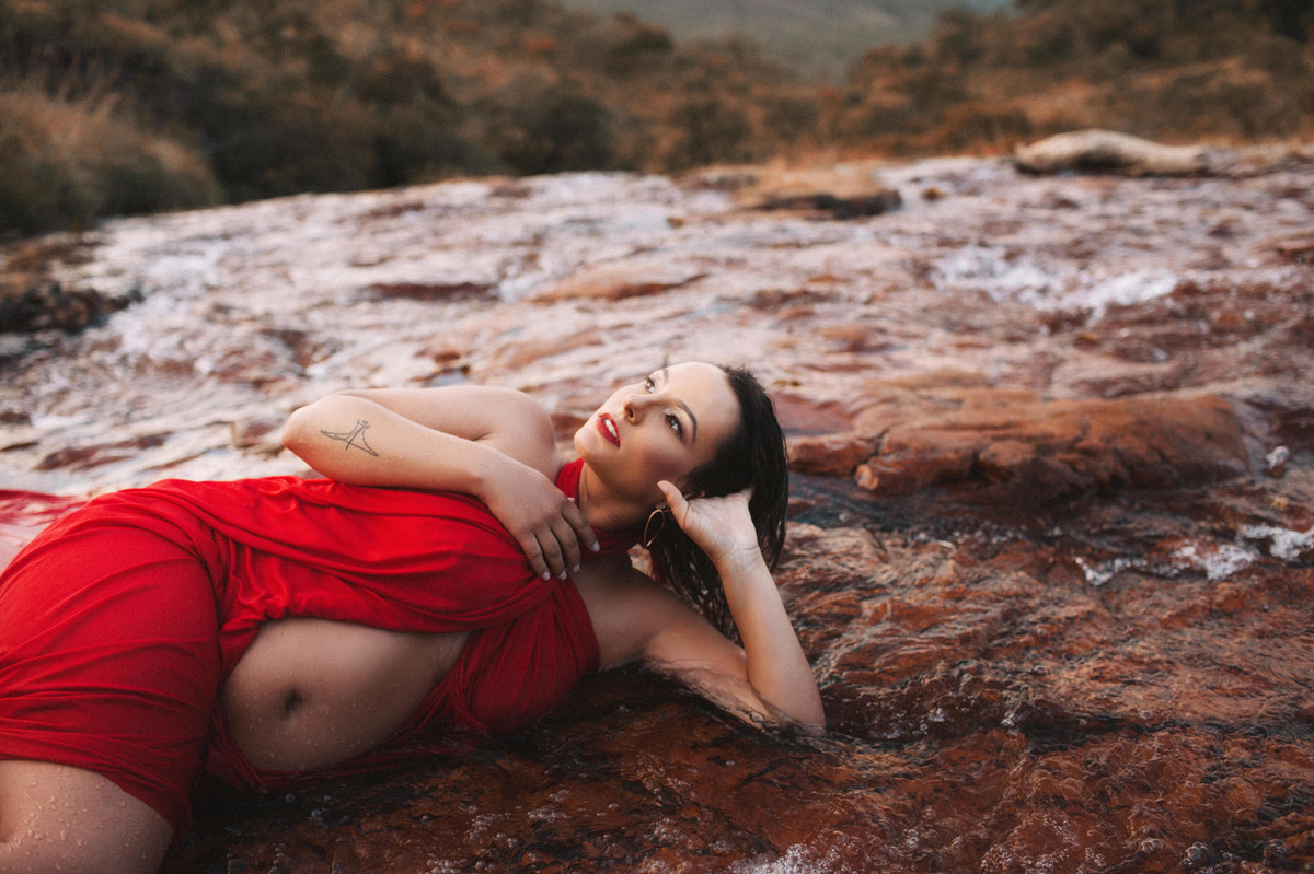 Grávida posando relaxada nas pedras do rio usando vestido vermelho olhando para as nuvens. Clicado pelo fotógrafo de gestantes Rafael Ohana em Brasília-DF