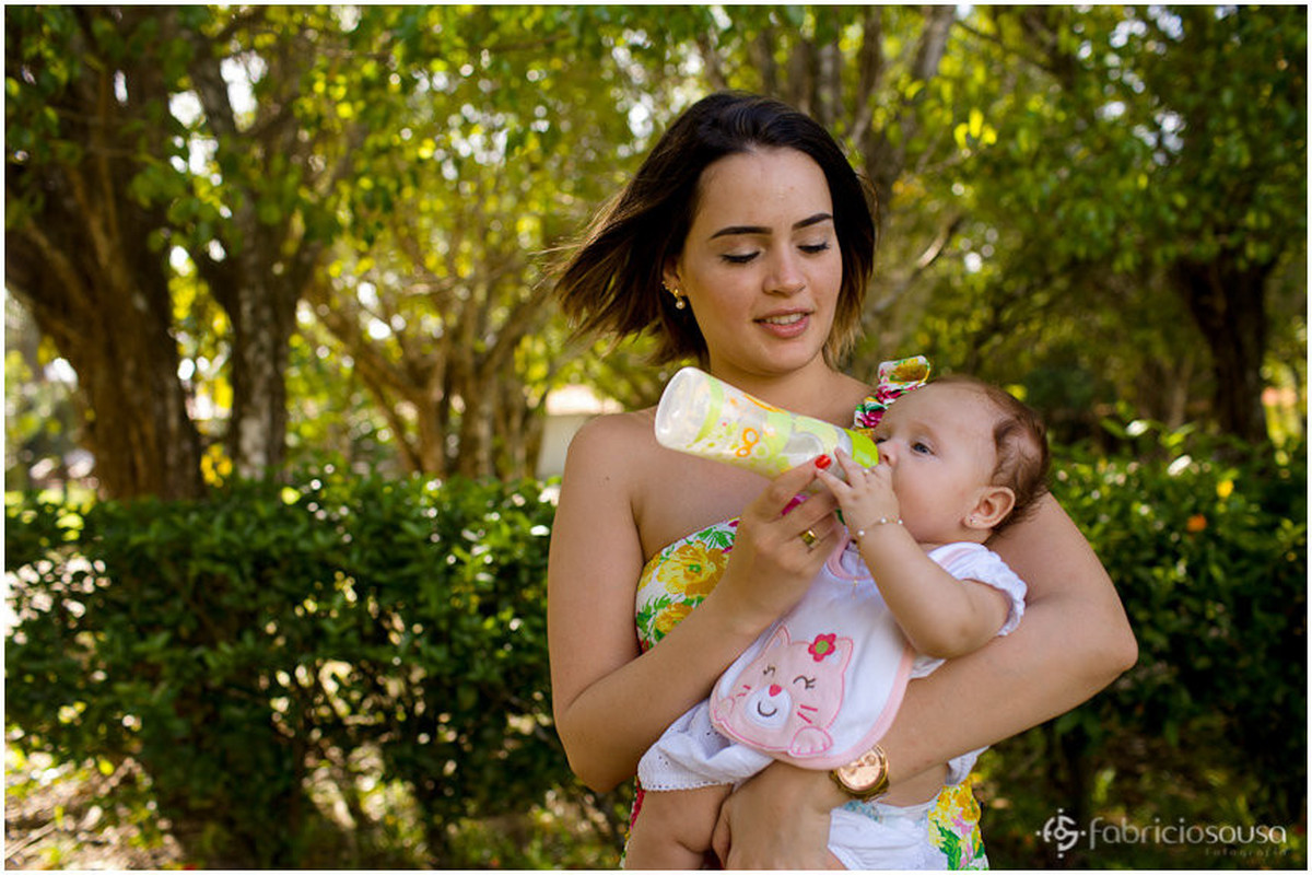 Mãe dando mamadeira para a bebê em seu colo em meio à natureza do parque