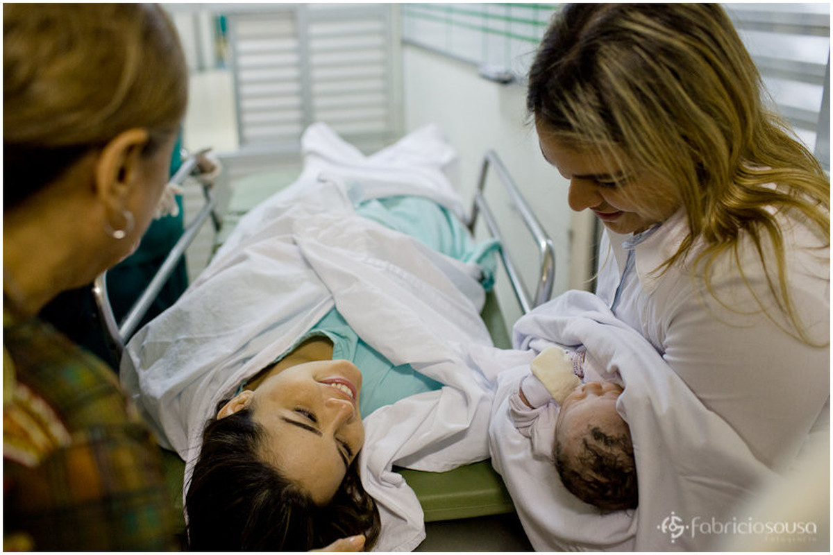 Na maca, mamãe contente observa a filha no colo da tia