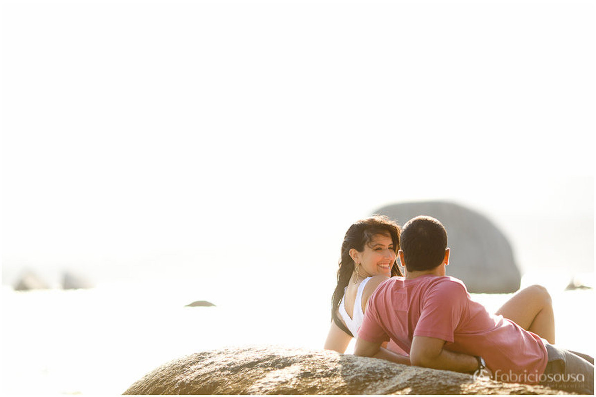 Casal de noivos aproveitam o sol escorados sobre uma pedra na beira do mar