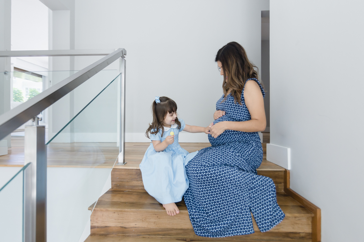 Mulher grávida sentada na escada de casa com a filha fazendo carinho na barriga