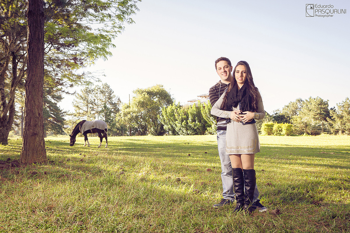 Em pasto, cavalo pastando e casal posando para foto em ensaio Pré-Wedding. Fotografia de Eduardo Pasqualini, fotógrafo de casamentos e ensaios em Rio do Sul, Santa Catarina. Lontras.