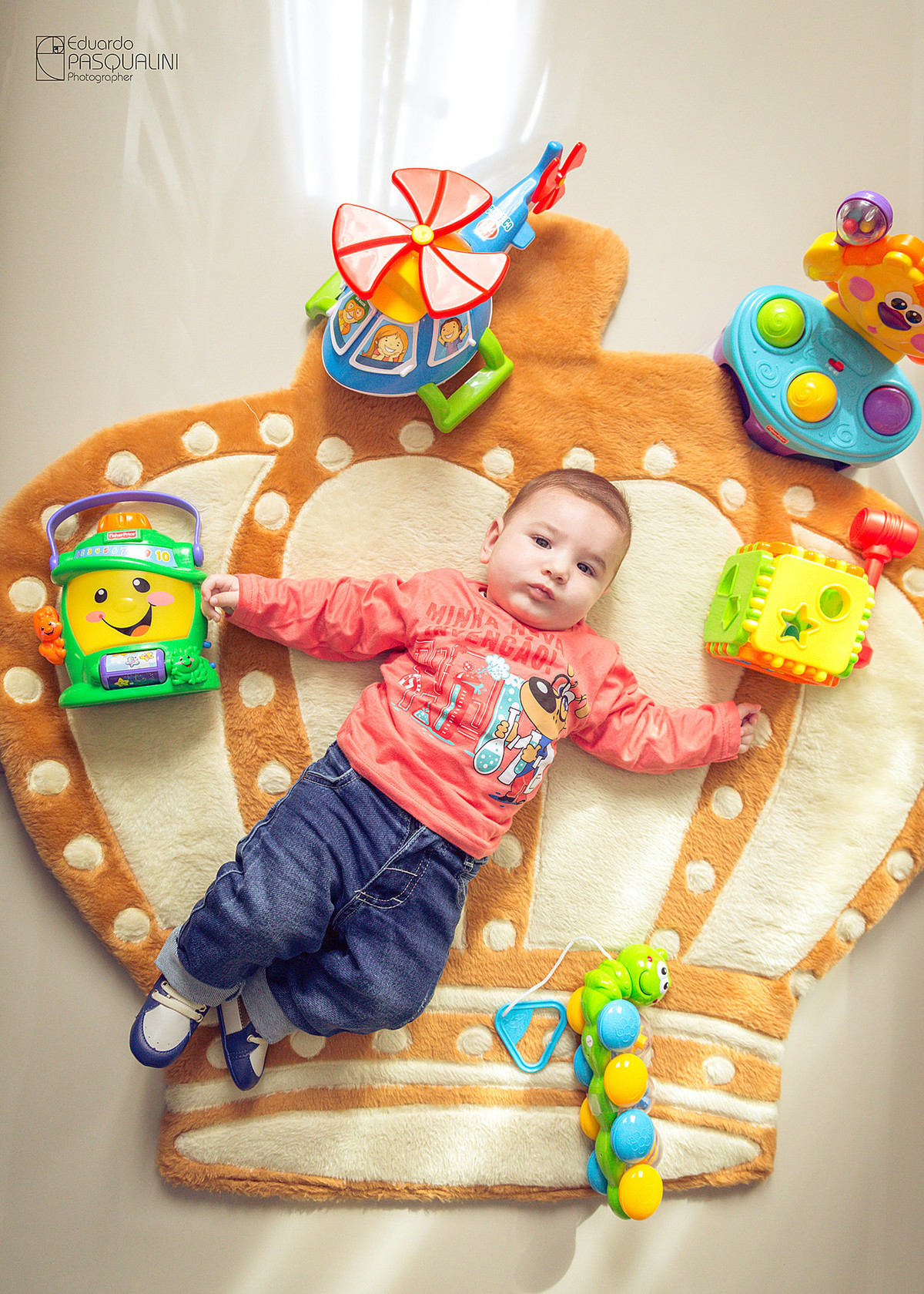 Foto de cima de criança em tapete de coroa de rei junto de seus brinquedos. Fotografia de Eduardo Pasqualini, fotógrafo de casamentos e ensaios em Rio do Sul, Santa Catarina.