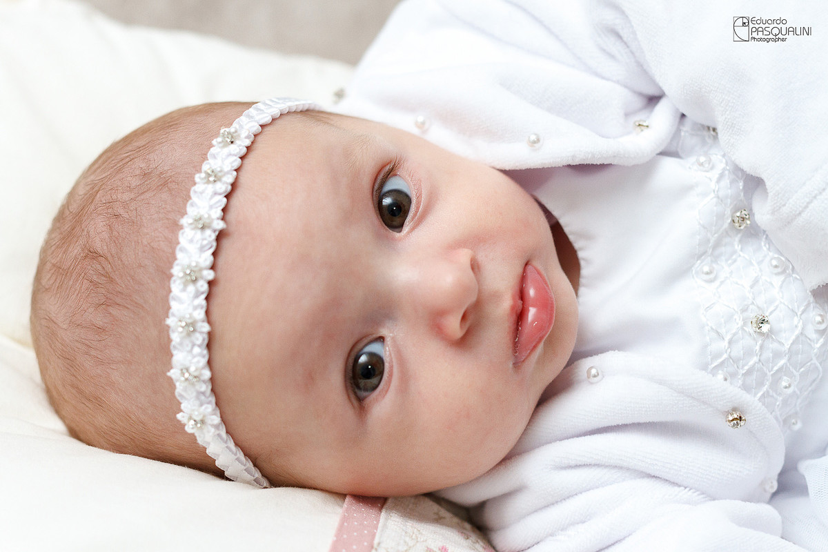 Olhar fixo de Alícia para foto com vestido de batizado. Fotografia de Eduardo Pasqualini, fotógrafo de casamentos e ensaios em Rio do Sul, Santa Catarina.