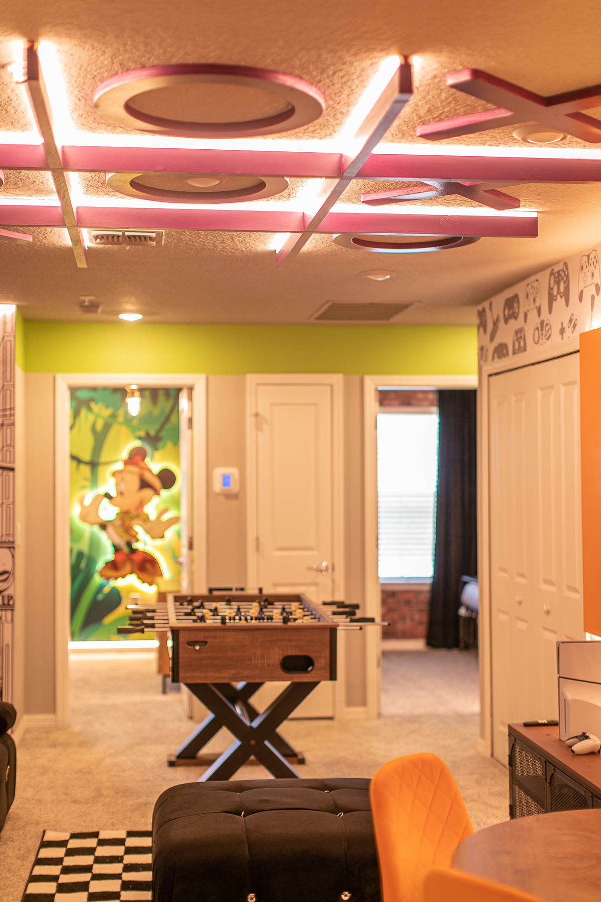 The image shows a vibrant game room with ceiling decor featuring XO shapes, a foosball table, and a mural of Mickey Mouse in the background. Real Estate and Interior Photography by Magic Lens Photo Media.