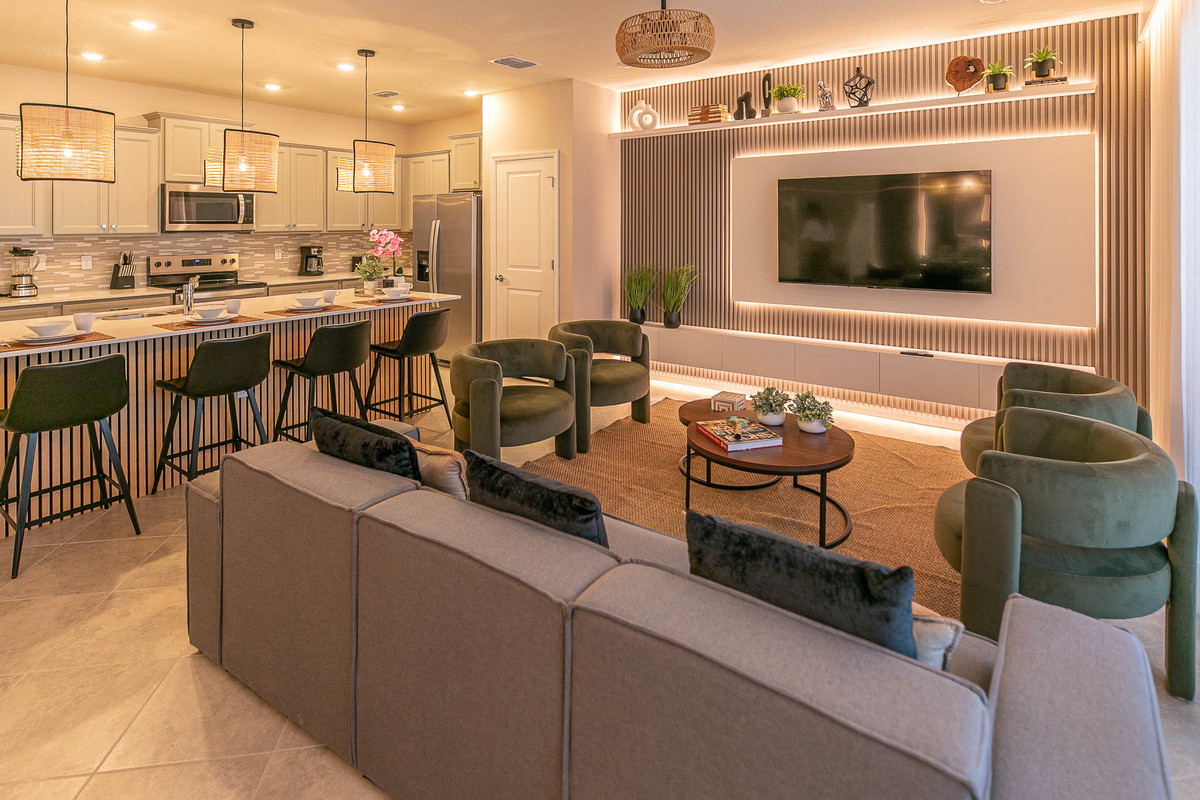 The image depicts a modern open-plan kitchen and living room with warm lighting, stylish seating, and a wall-mounted TV surrounded by shelves and decor items. Real Estate and Interior Photography by Magic Lens Photo Media.