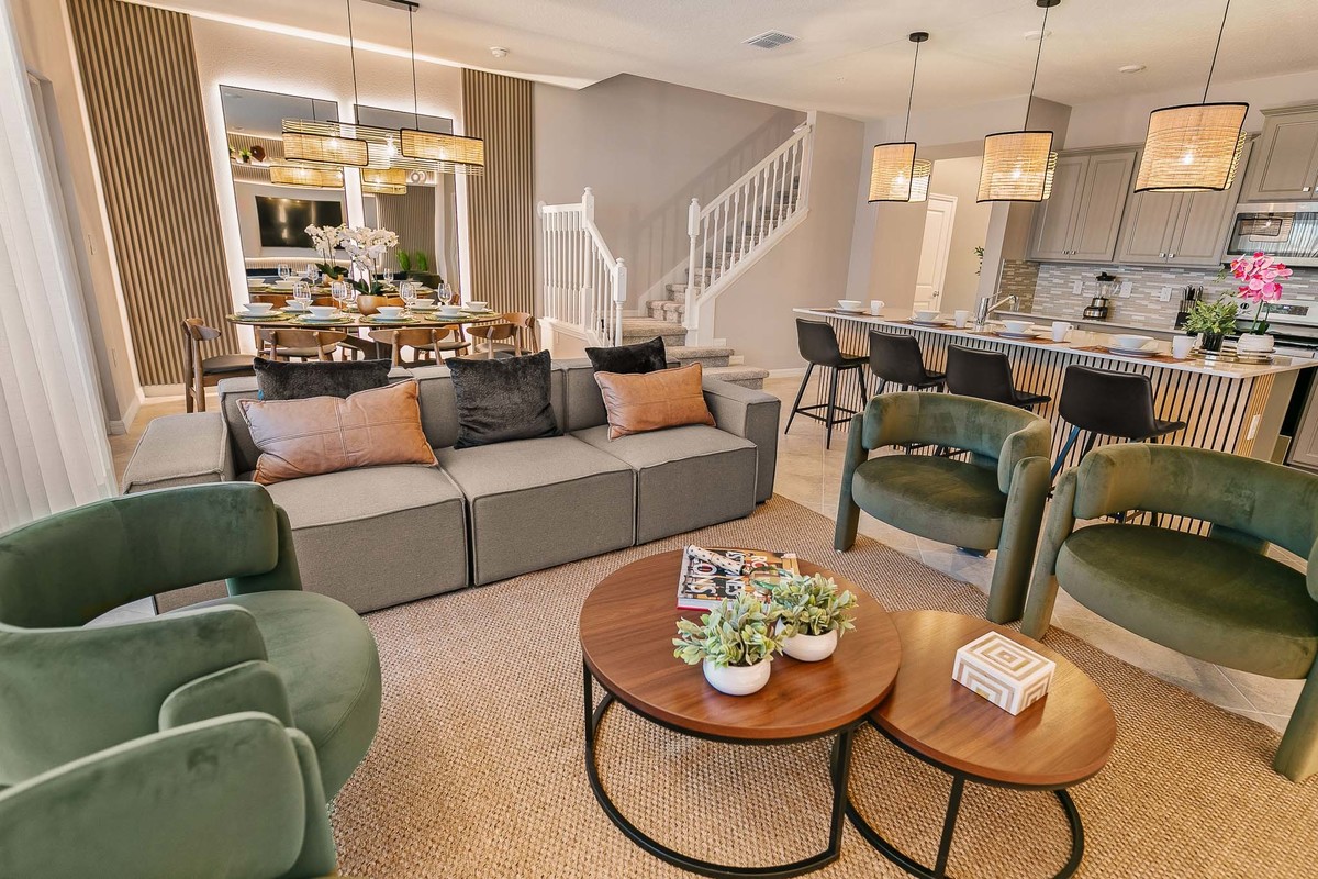 Open-plan living area with a grey sectional sofa, green armchairs, and wooden tables. Kitchen bar in the background features black stools and rattan pendant lights. Real Estate and Interior Photography by Magic Lens Photo Media.