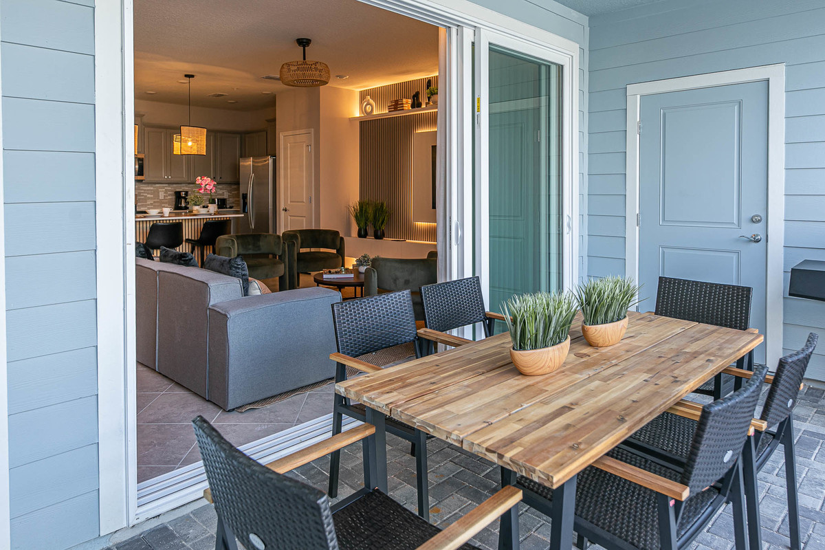 Outdoor dining area with a rustic wooden table, black chairs, and potted plants, overlooking the living room. Real Estate and Interior Photography by Magic Lens Photo Media.