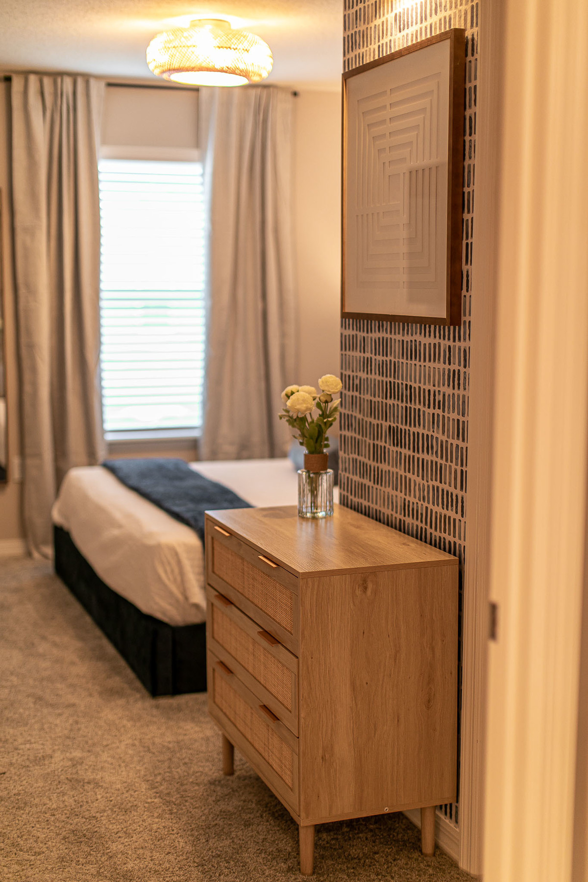 The image shows a cozy bedroom with a wooden dresser adorned with flowers in a glass vase. A patterned wall holds a geometric framed art piece, and soft natural light filters through a window. Real Estate and Interior Photography by Magic Lens Photo Media