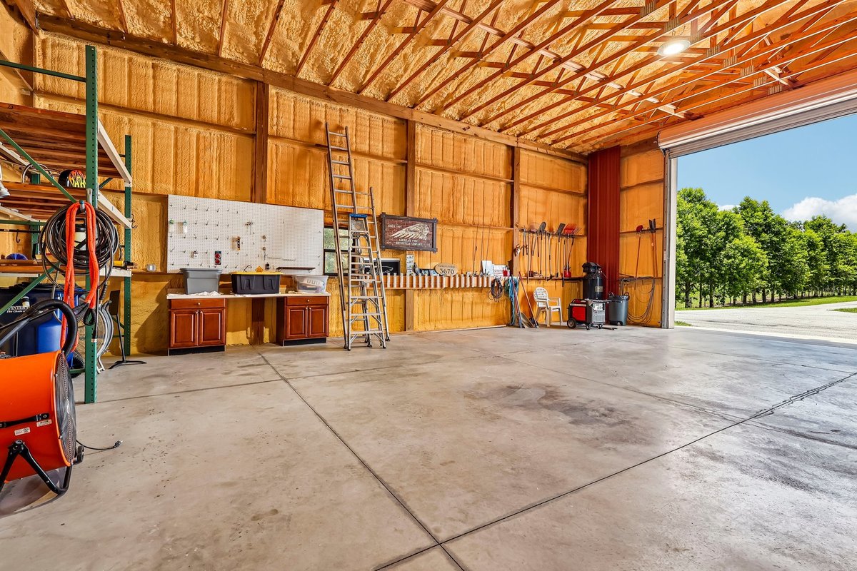 Wide interior view of a spacious insulated workshop with high ceilings, tool storage, concrete flooring, and natural light from the open bay door.