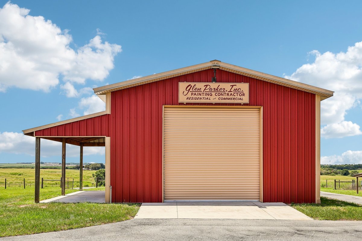 Front view of a red metal workshop barn with a large roll-up door, covered side area, driveway, and open rural land in Haines City, Florida.
