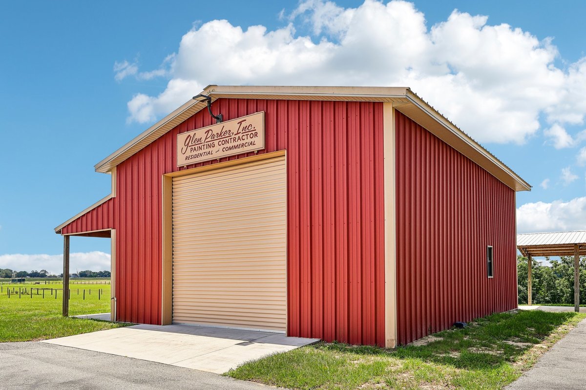 Angled exterior view of a red metal workshop barn with roll-up garage door, side entrance, and open acreage surrounding the property.
