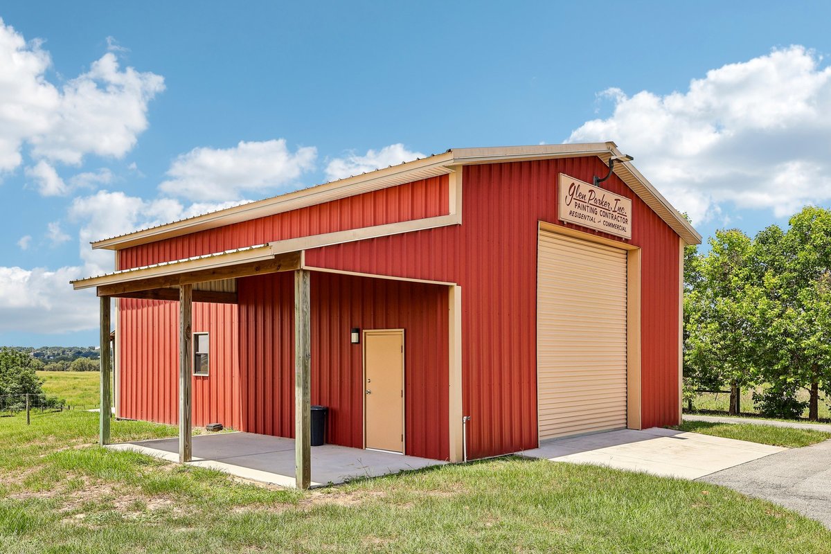 Side exterior view of a red workshop barn with covered porch, roll-up door, paved driveway, and open pasture views.