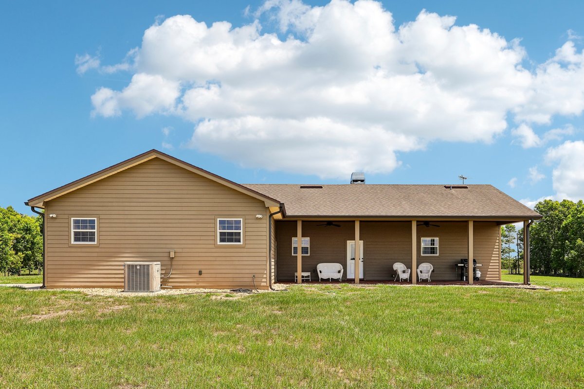 Rear exterior view of a single-story rural home with covered patio, seating area, open lawn, and countryside views.
