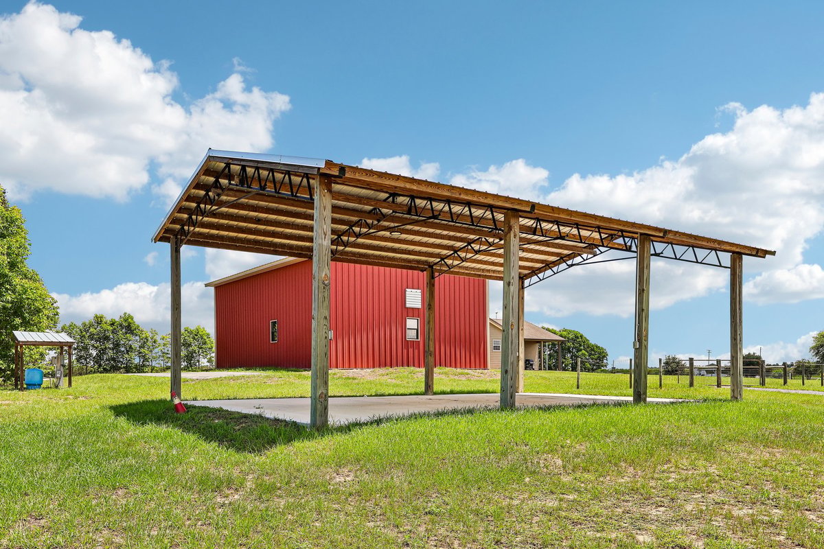 Covered outdoor structure beside a red workshop barn, offering flexible space for equipment, parking, storage, or rural property use.