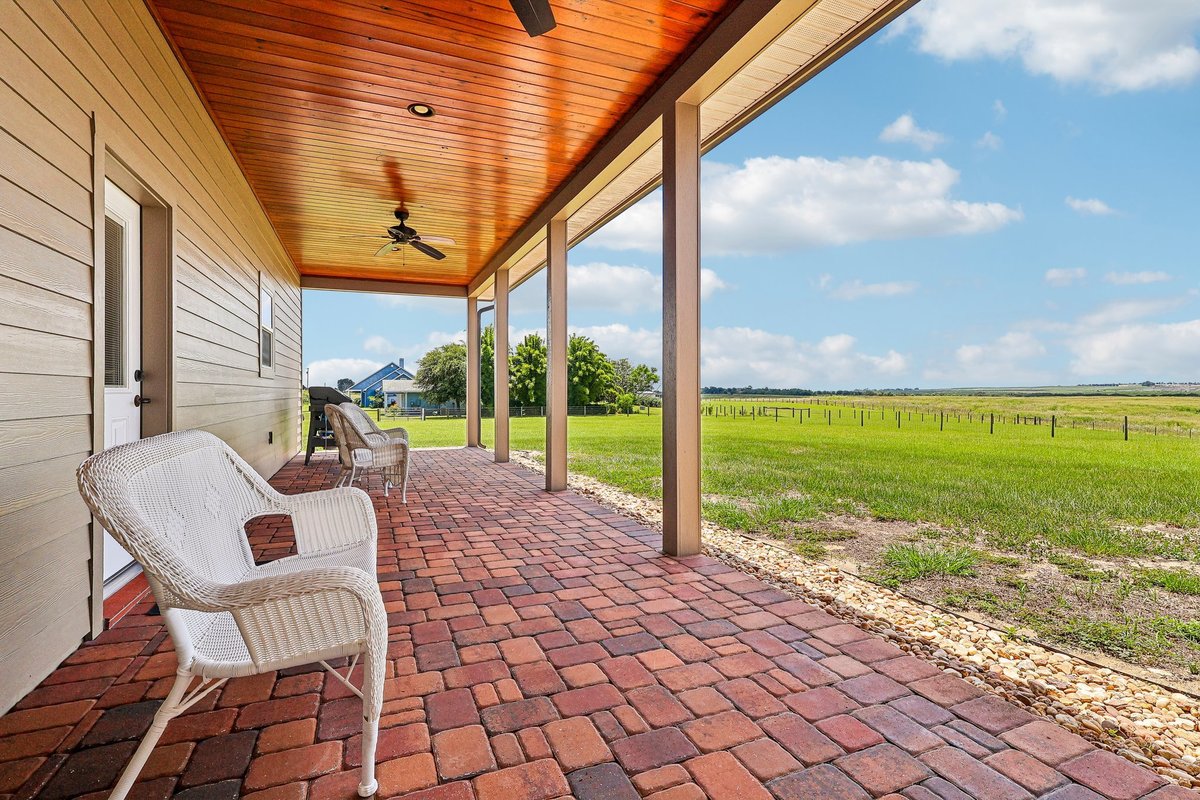 Covered back porch with brick pavers, white seating, wood ceiling, ceiling fan, and open views across the rural acreage.
