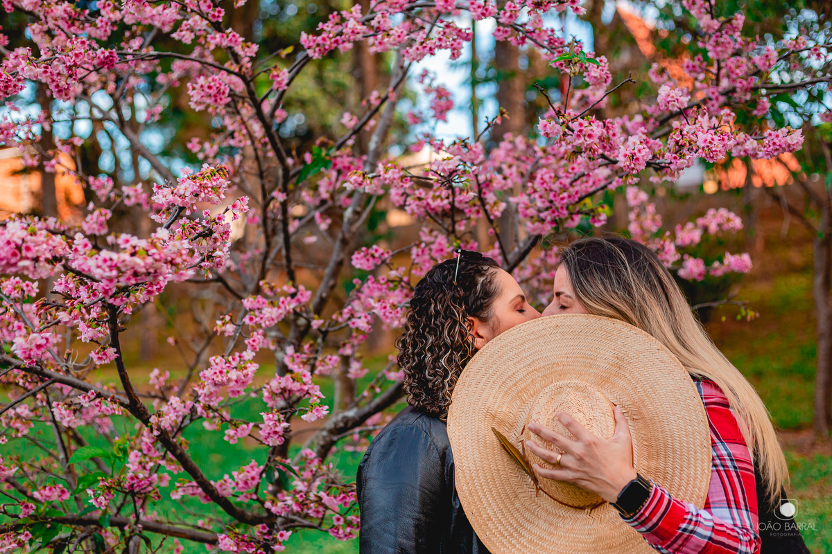 casal gay de mulheres se beija escondendo o rosto com chapéu de palha, com cerejeira no fundo