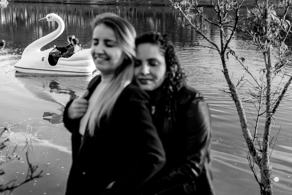 foto preto e branco casal gay de mulheres se abraçando de olhos fechados com pedalinho em forma de cisne ao fundo no lago