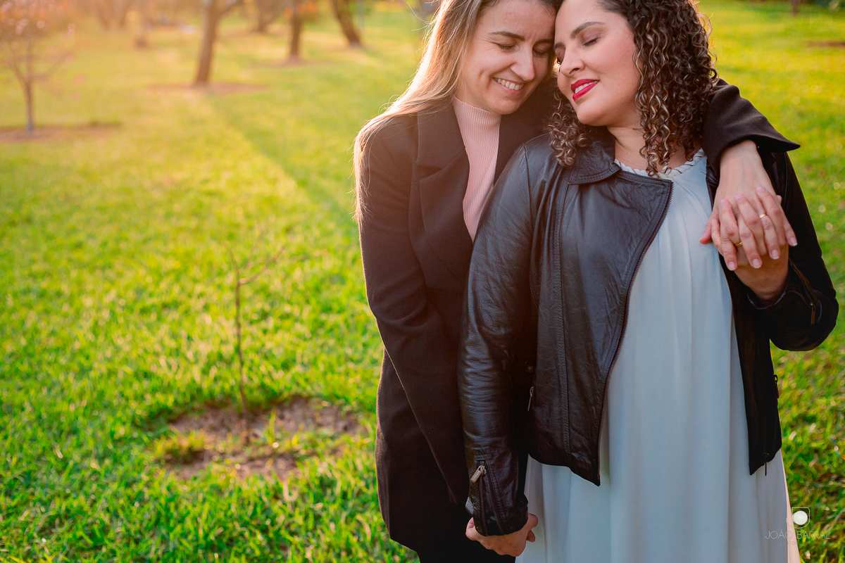 casal gay de mulheres se abraçando de olhos fechados em jardim com luz do sol e árvores no fundo