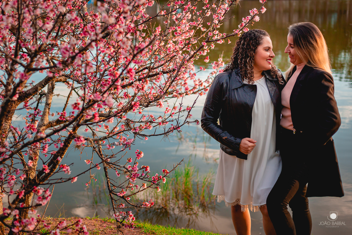 casal gay de mulheres abraçadas se olhando com lago e cerejeira ao fundo