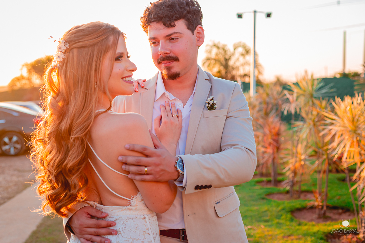 casal de noivos abraçados em jardim com por do sol e noiva olhando para o lado