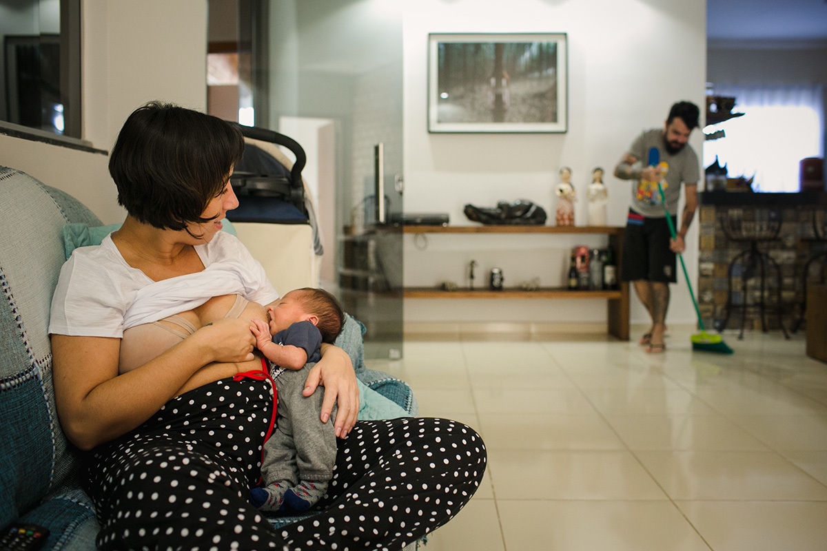 foto feita por adrianacostafotos do pai varrendo a casa enquanto a mãe amamenta