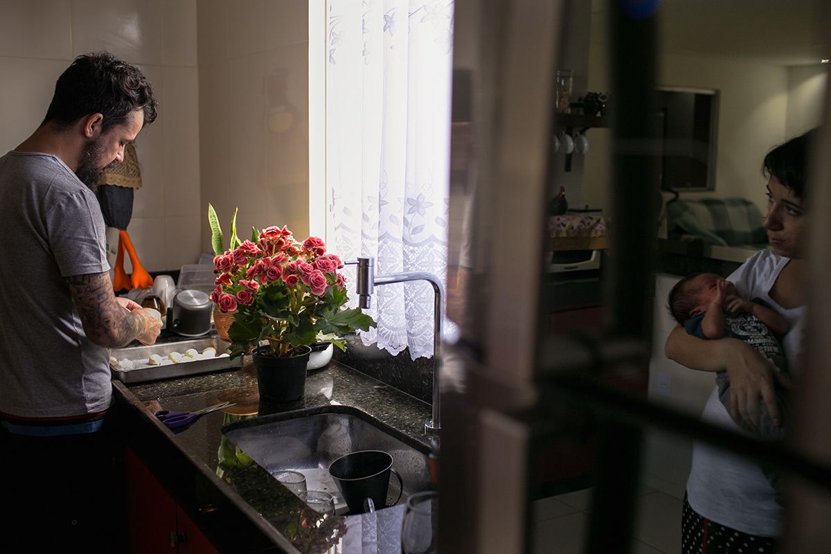 reflexo da mãe com o bebê no colo observando o pai na cozinha em uma fotografia feita por Adriana Costa