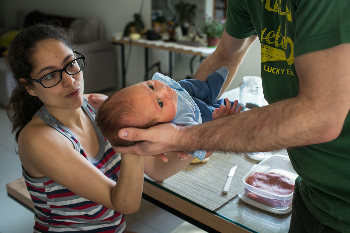 pai passando seu bebê para o colo da mãe, em uma foto feita por Adriana Costa