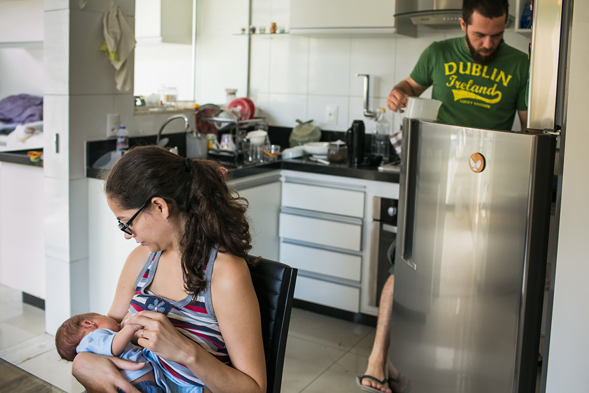 pai organizando a cozinha enquanto a mãe amamenta seu bebê em foto de Adriana Costa