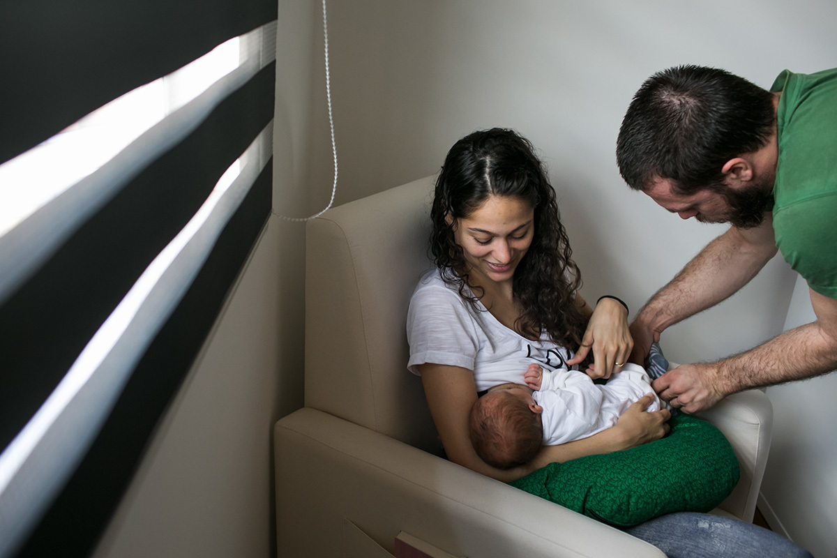 Foto de Adriana Costa do pai colocando meia no seu bebê enquanto ele amamenta