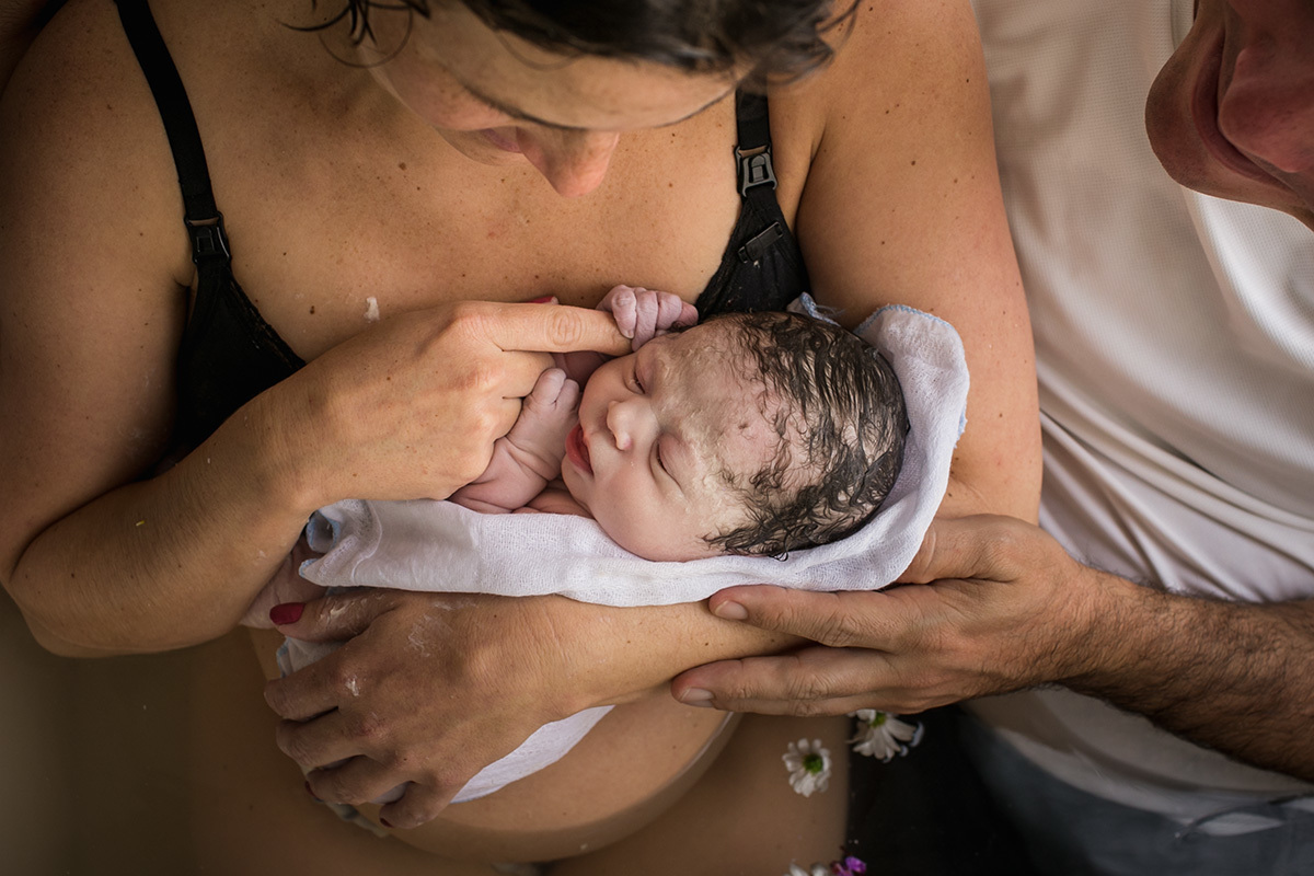 foto de Adriana Costa do bebê segurando o dedo da mãe logo após nascer, com o pai ao lado