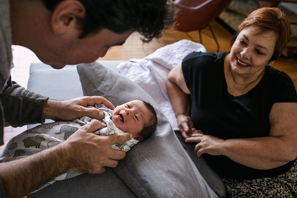 bebê rindo para o pai e avó sorrindo ao ver a cena, em foto de Adriana Costa