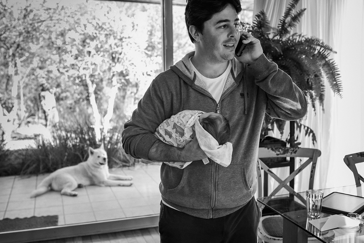 pai falando no celular e com a outra mão carregando sua pequena bebê recém-nascida, por Adriana Costa