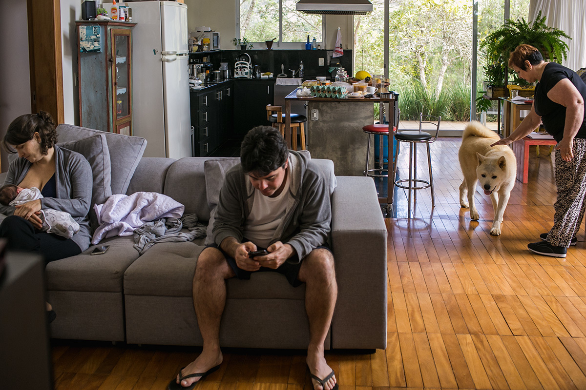 mãe amamentando enquanto o pai vê o celular e a avó brinca com o cão da casa, por Adriana Costa