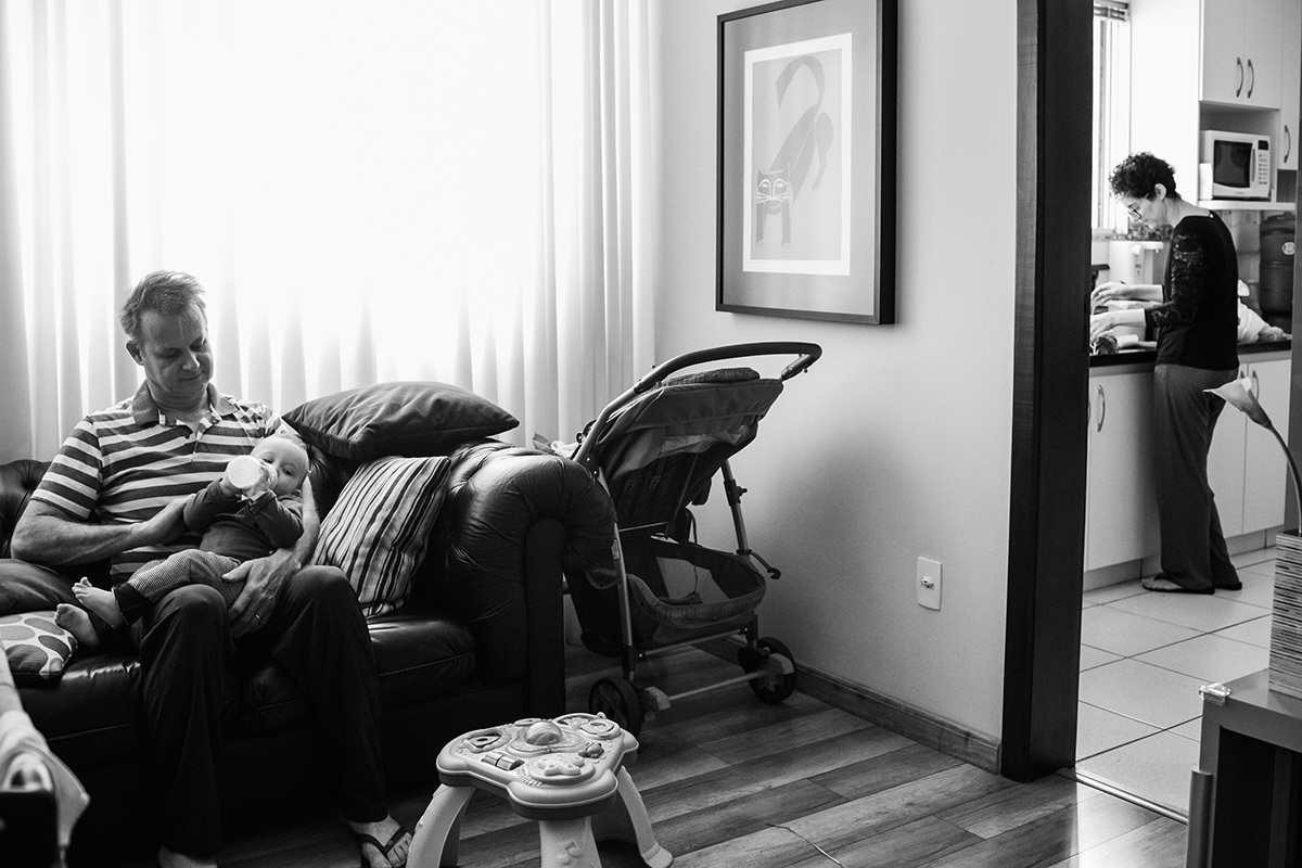 pai dando mamadeira ao filho enquanto mãe está na cozinha, por Adriana Costa