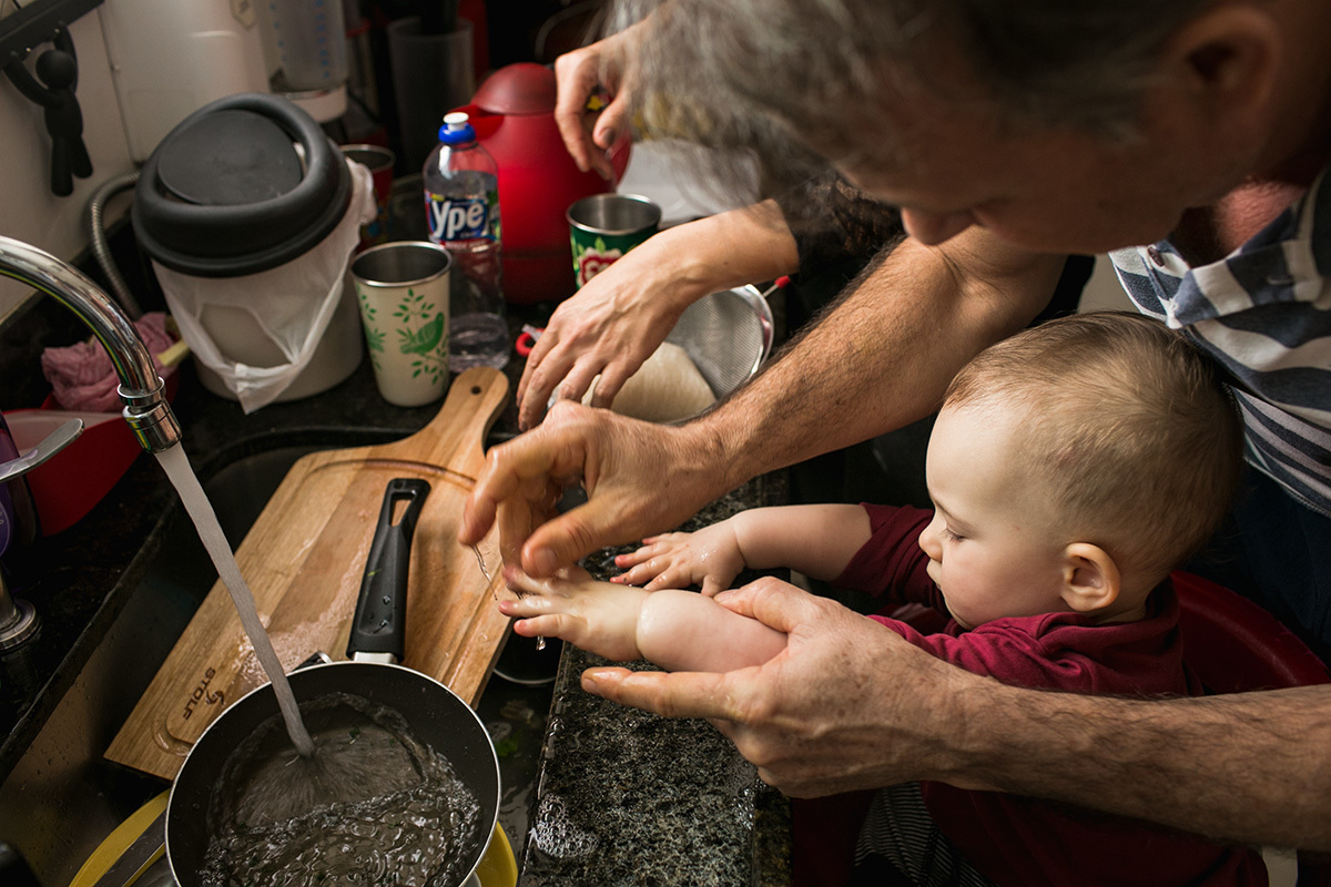 pai lavando as mãos do seu bebê na pia da cozinha que está cheia de vasilhas, em foto de Adriana Costa