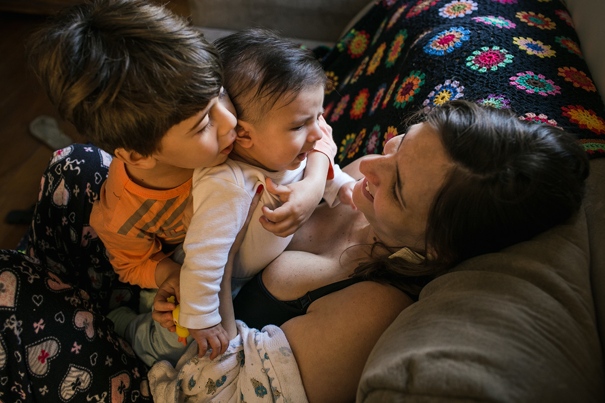 irmão mais velho abraçando o irmão caçula que está no colo da mãe, por adrianacostafotos
