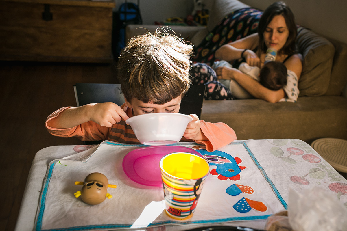menino tomando leite no pires enquanto sua mãe da mamadeira para seu irmão em foto de Adriana Costa