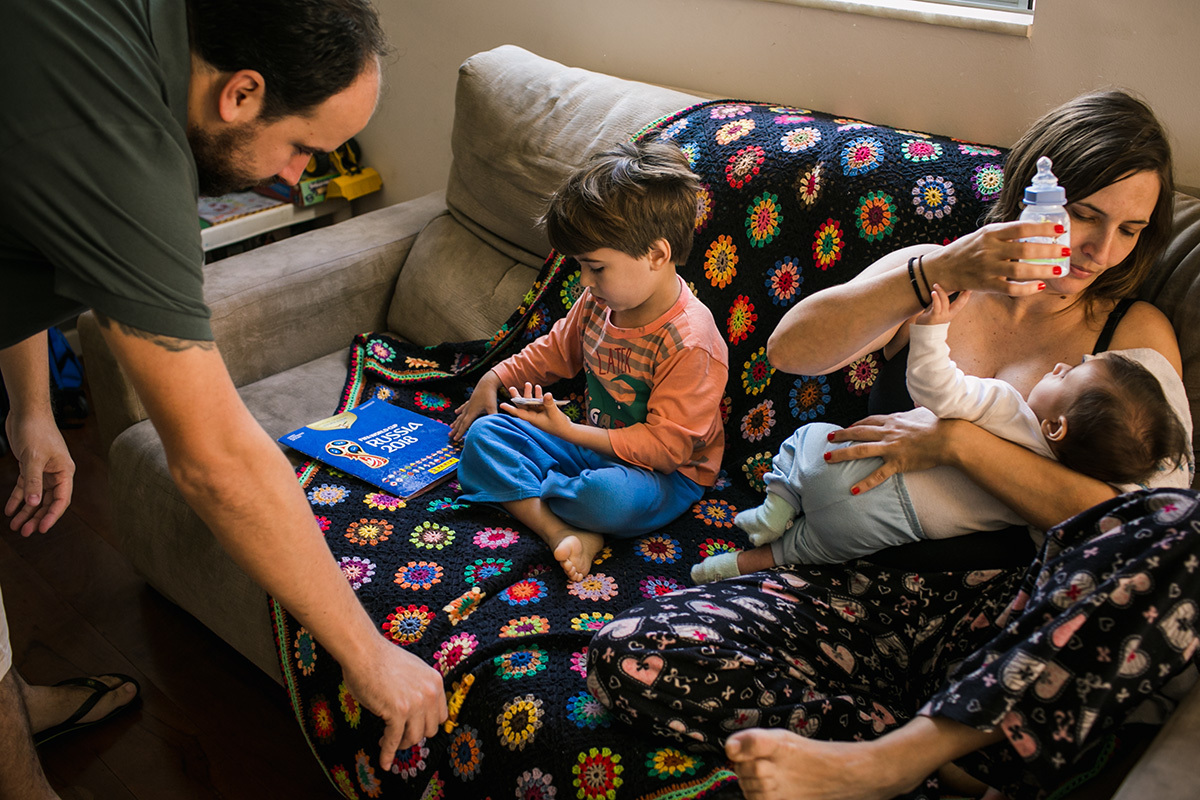 família reunida no sofá, pai e filho álbum de figurinhas e mãe dando mamadeira para o bebê, por adrianacostafotos