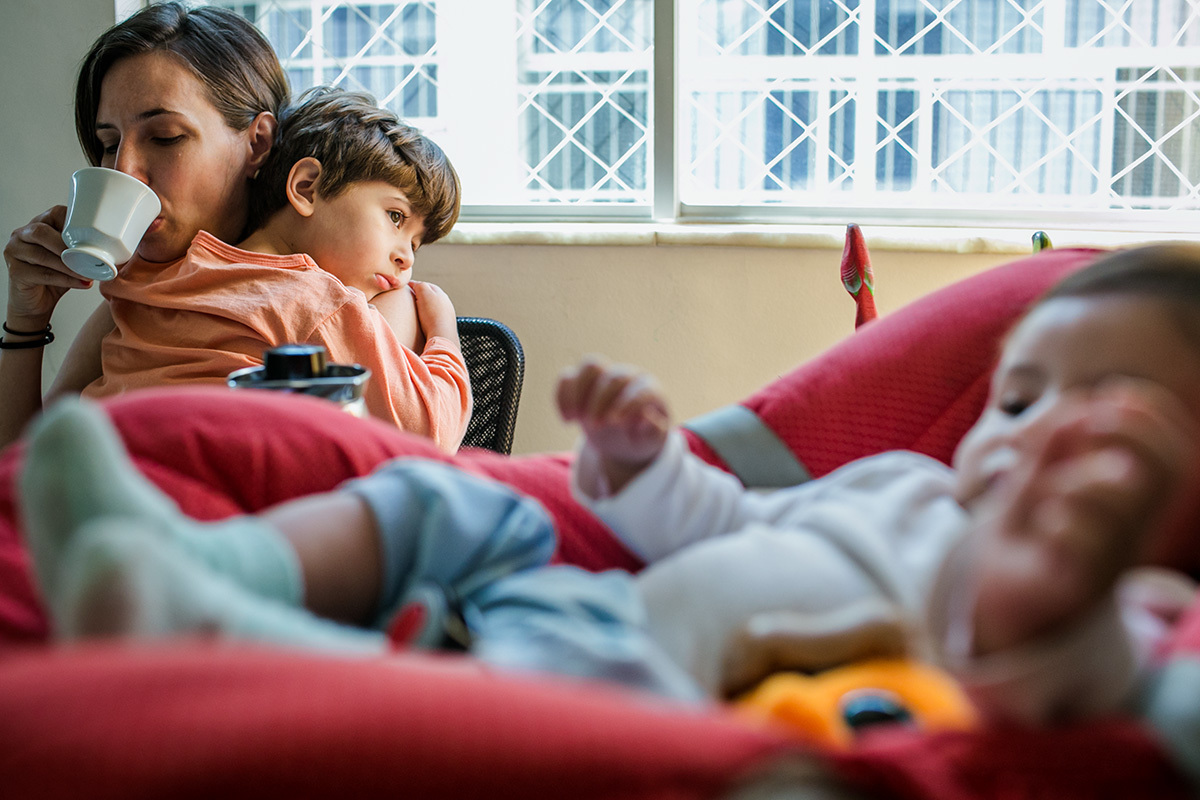 mãe tomando seu café com seu filho triste no colo e seu bebê sobre a mesa no bebê conforto, por adrianacostafotos