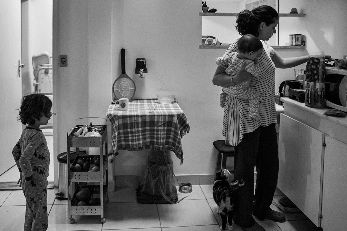 mãe com bebê no colo e seu filho mais velho preparando o café da manhã, por adrianacostafotos