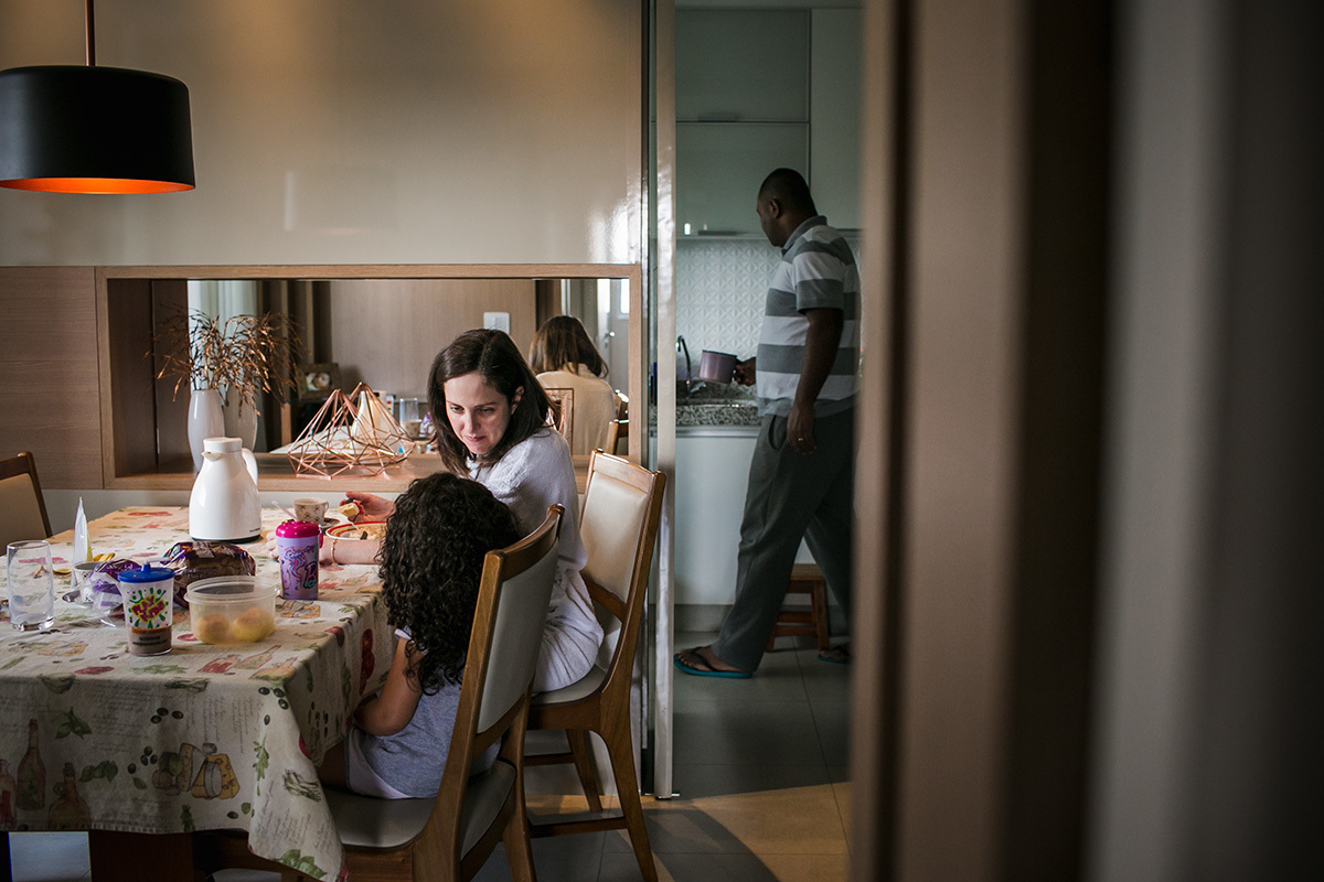 mãe e filha tomando café enquanto o pai prepara o leite na cozinha, em foto de Adriana Costa