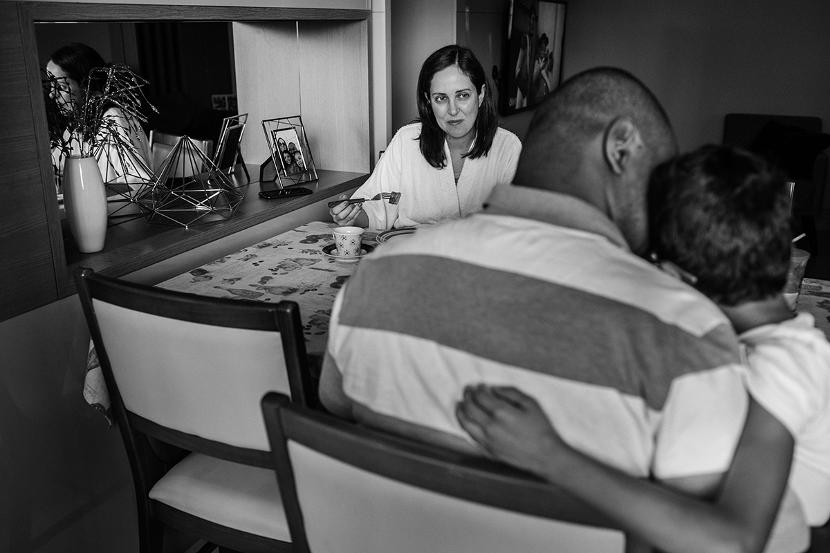 mãe observa marido e filho se abraçarem no café da manhã, em foto de Adriana Costa