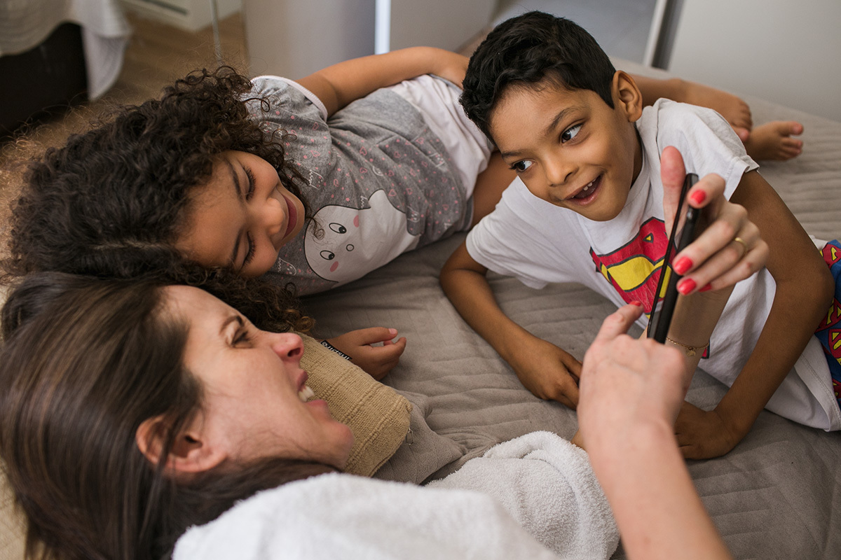 mãe brincando com seus filhos na cama, em foto de Adriana Costa
