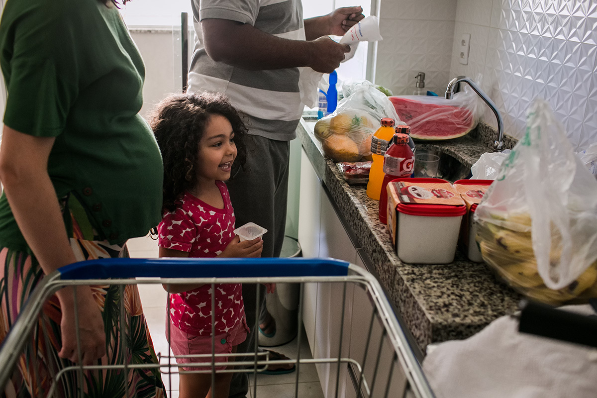 filha vendo as compras de supermercado com os pais na cozinha de casa, em foto de Adriana Costa
