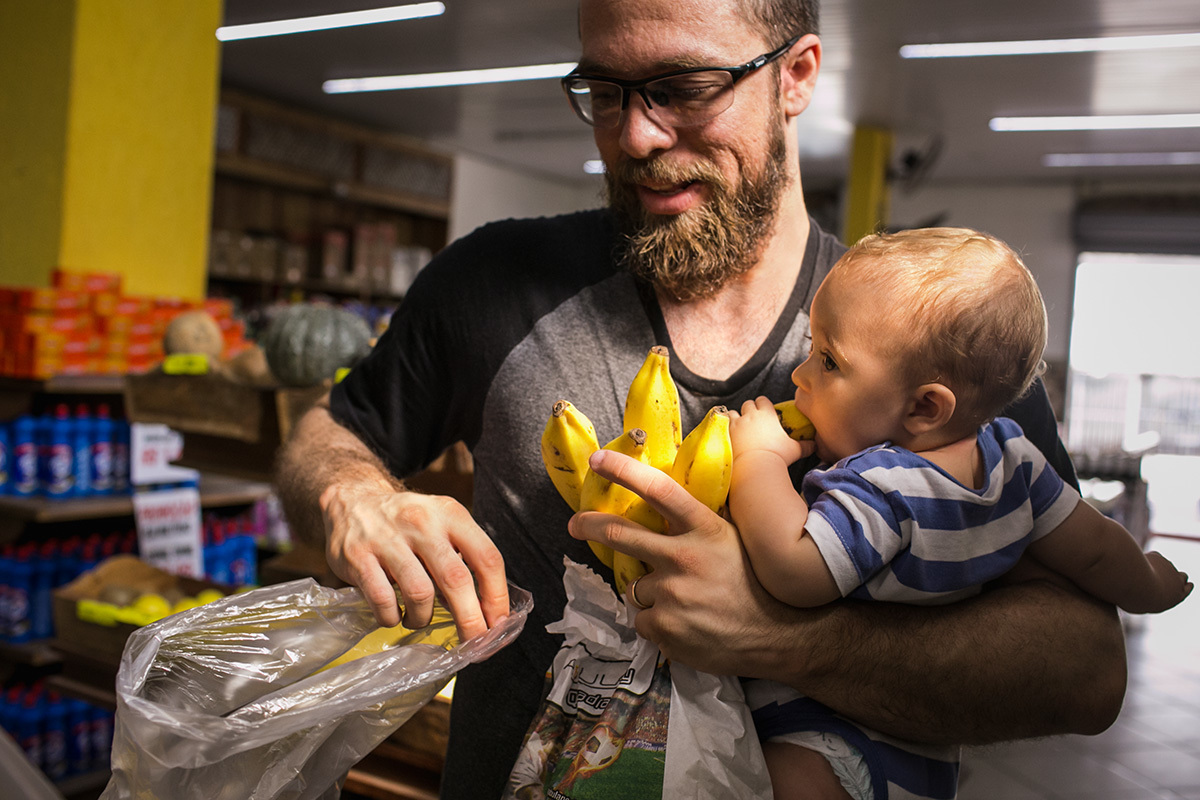 pai escolhendo a banana para comprar e seu bebê colocando-a na boca sem ele ver, em foto de Adriana Costa