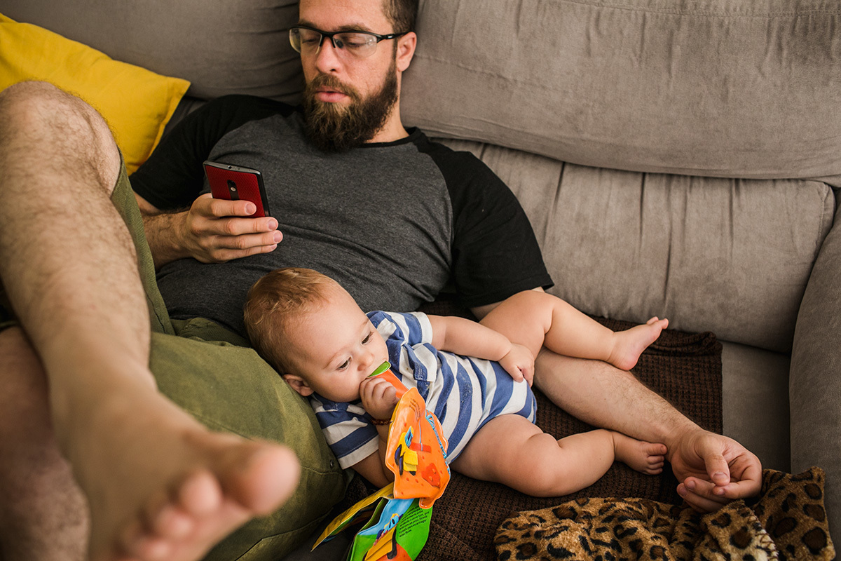 pai vendo o celular enquanto seu bebê brinca no seu colo, por adrianacostafotos