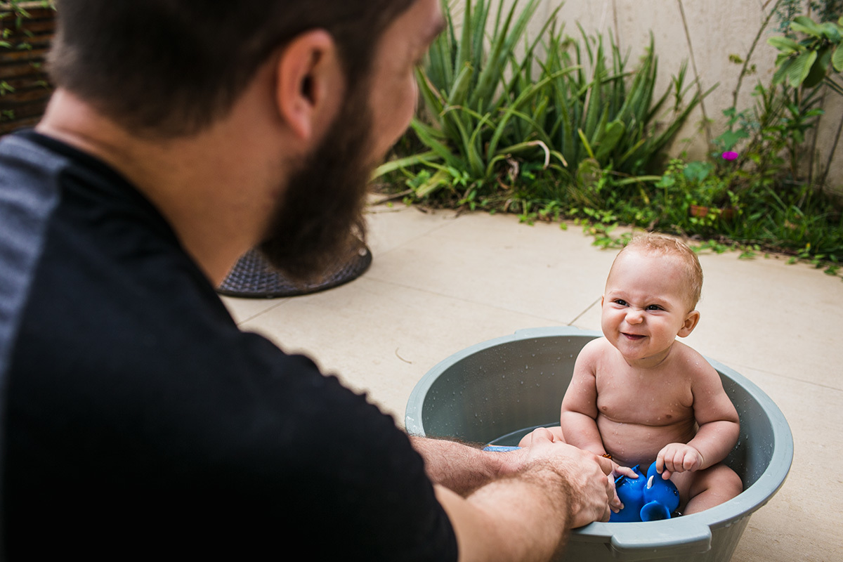 bebê feliz fazendo caretinha para o pai durante o banho de bacia no quintal de sua casa, por adrianacostafotos